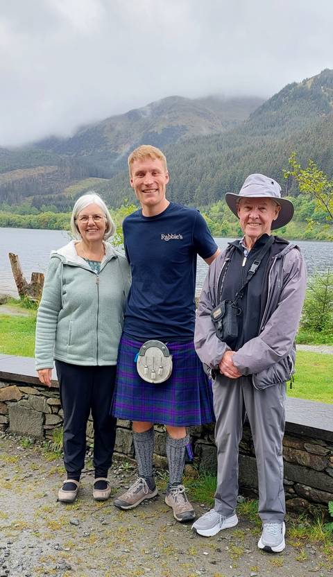 Three travellers posing in front of a lakeside backdrop, one wearing a kilt and two older companions smiling.