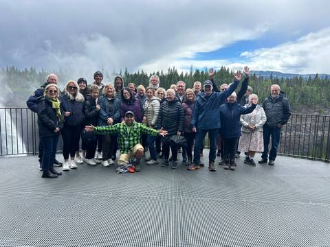A large tour group in outdoor gear posing happily on a viewing platform beside a misty waterfall and forest.