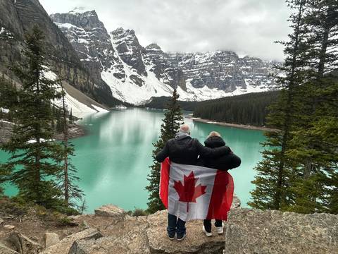 A couple wrapped in a Canadian flag gazing over the turquoise waters of Moraine Lake amid snow-capped peaks and evergreens.