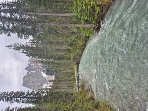 A fast-flowing alpine river flanked by evergreens with a jagged snow-dusted peak rising beyond.