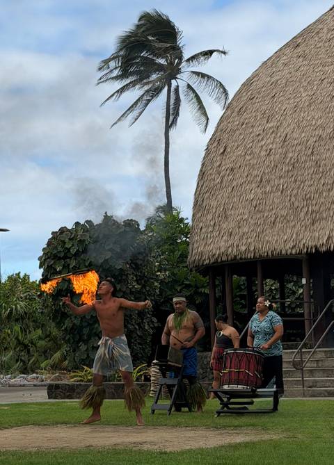 Fire dancer performs in front of thatched buildings while others watch
