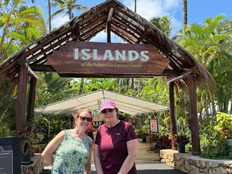 Two women smiling under a wooden sign reading 'Islands of the Polynesian Cultural Center'