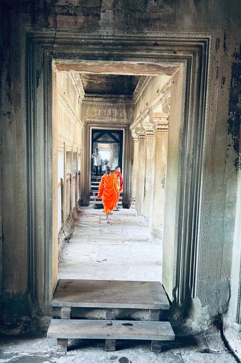 Buddhist monks in bright orange robes walk through a stone corridor of an ancient temple with detailed carvings.