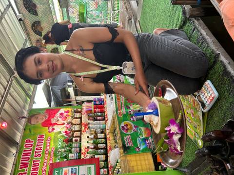 Smiling young woman kneels beside a tray with coconut ice cream and orchid garnish at a vibrant market stall.