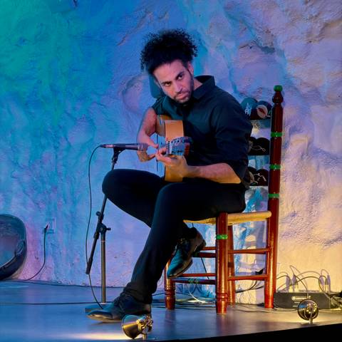 Flamenco guitarist seated on a wooden chair performing under blue stage light.