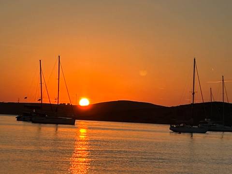 Brilliant orange sunset sets behind low hills while sailboats anchor on calm reflective sea.