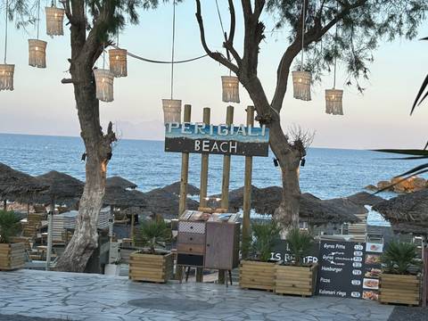 Beachfront restaurant sign reading 'Perigiali Beach' with thatched umbrellas and blue sea beyond.