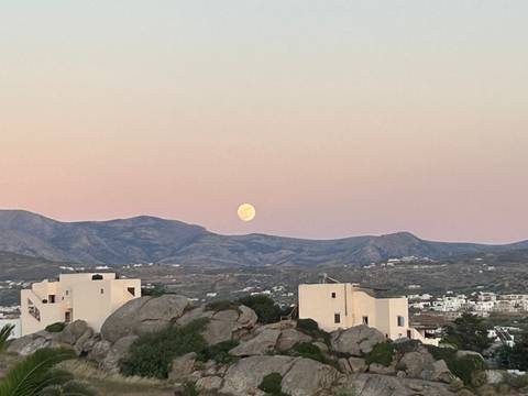 Rising full moon over pastel sky above Cycladic white houses and rocky hills.