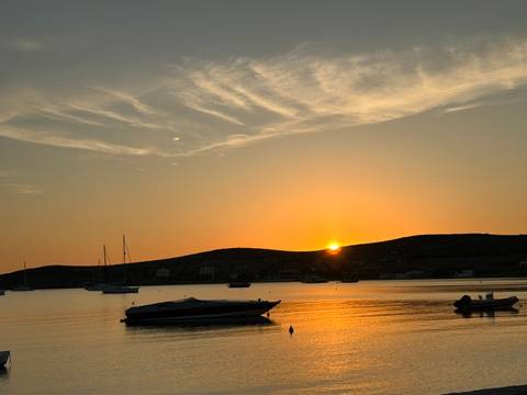 Sunset over a calm bay with small boats silhouetted against an orange sky and low rolling hills.