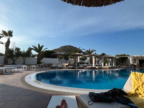 Resort swimming pool bordered by loungers, palms and a distant hill under a clear blue sky.