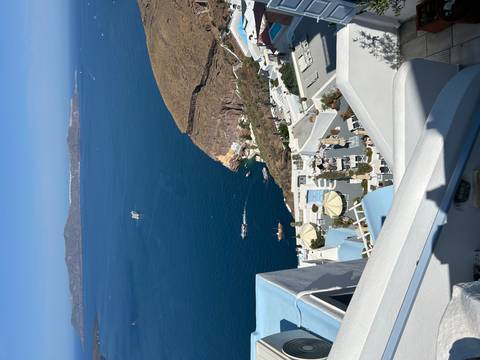 Cliff-top view of whitewashed villas cascading toward the deep blue Aegean with boats far below.