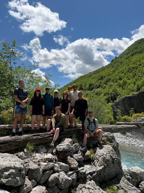 Group of hikers pose on a fallen log amid forested mountains under a partly cloudy sky