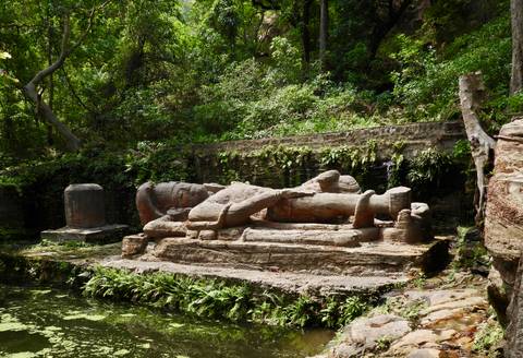Ancient stone sculpture of a reclining figure near a water pool in a lush forest setting.
