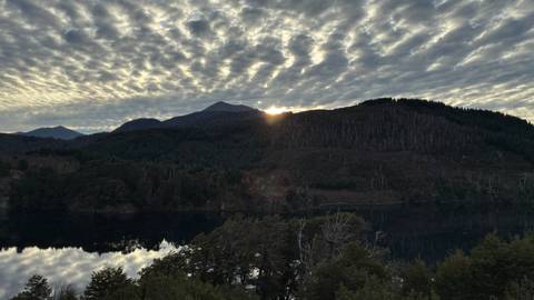 Morning sun peeking over a mountain ridge reflecting on a calm forest lake under patterned clouds.