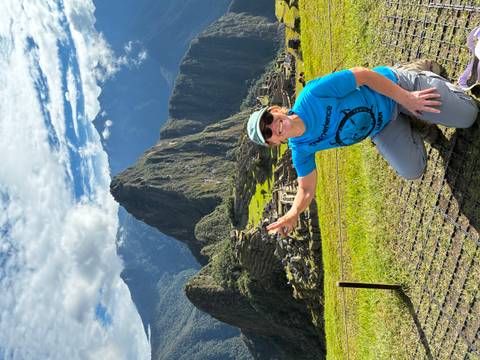 Traveler kneels and flashes a peace sign with Machu Picchu rising majestically behind.