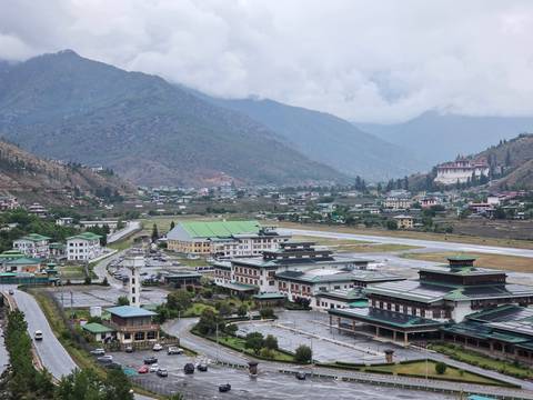 A small mountain airport runway with surrounding town and monastery set in a green valley under low clouds.
