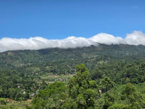 Verdant mountains rise above a valley town as low clouds drift along the ridgeline under a clear blue sky.