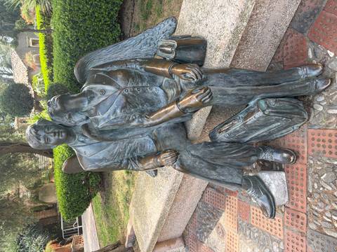 Bronze sculpture of a suited man and woman with wings resting together on a stone bench in a shaded garden