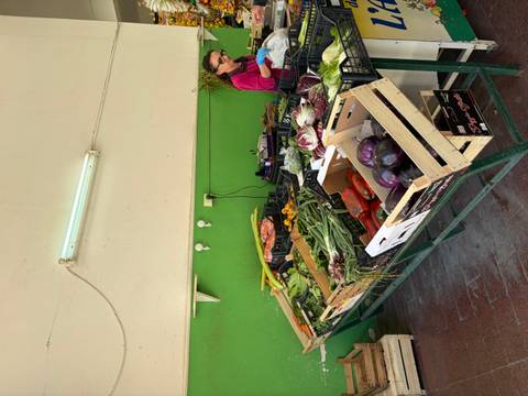 Small green-painted produce stall displaying fresh vegetables with a vendor weighing goods behind the counter