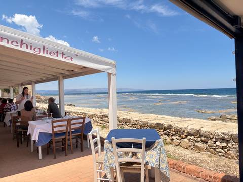Open-air seaside restaurant with diners enjoying lunch beside the rocky coastline and turquoise sea