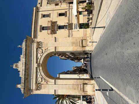 Grand stone triumphal arch leading into a historic street lined with baroque buildings under clear skies
