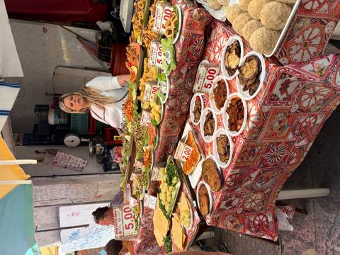 Abundant market stall of prepared Sicilian dishes and a vendor standing behind price signs