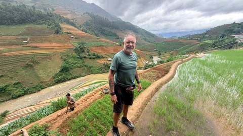Smiling hiker stands on a narrow rice-terrace path high above a green valley dotted with fields and misty mountains