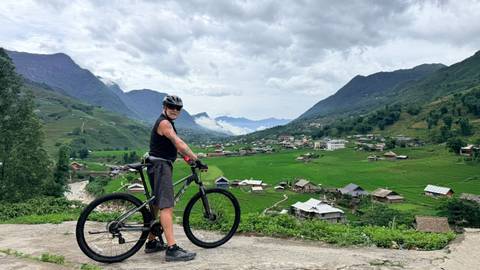 Cyclist poses with mountain bike overlooking a lush valley of rice fields and distant peaks under cloudy skies