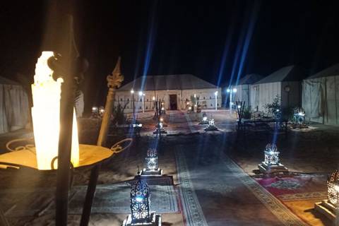 Nighttime desert camp illuminated by lanterns and rugs leading to canvas tents.