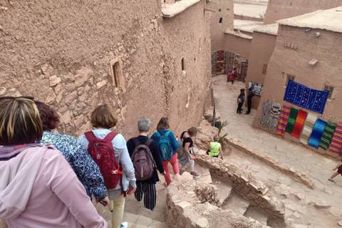 Tourists descend narrow stone steps between adobe walls lined with colorful textiles.