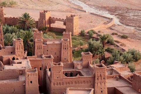 Golden light bathes the earthen towers and palm trees of Ait Benhaddou beside a dry river.