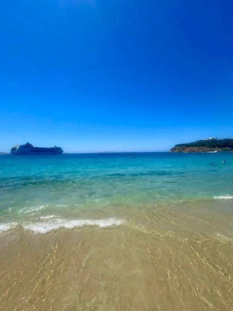 Turquoise waters lap a sandy Greek island beach while a large cruise ship anchors offshore under a bright blue sky.