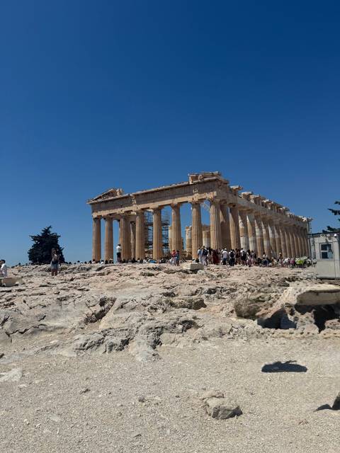 The Parthenon stands majestic on the Acropolis as crowds explore the ancient marble ruins against a deep blue sky.