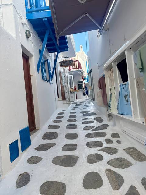 Narrow cobblestone alley with bright blue shutters and boutique shops winds through the white Cycladic architecture.