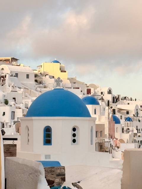 Iconic whitewashed buildings topped with vivid blue domes crowd the cliffs of Oia in soft evening light.