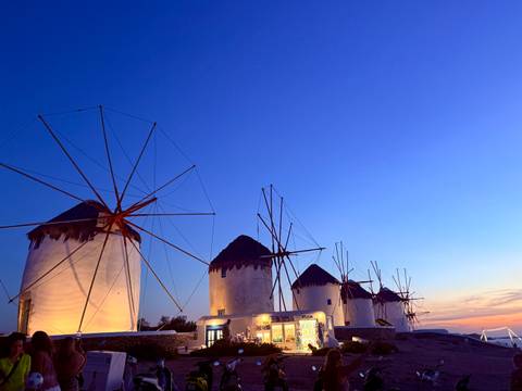 The traditional windmills of Mykonos stand against a deepening twilight sky with warm lights below.