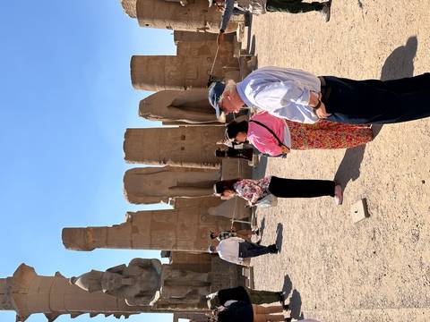 Tourists observing fragmented colossal statues in open-air temple courtyard under bright sky