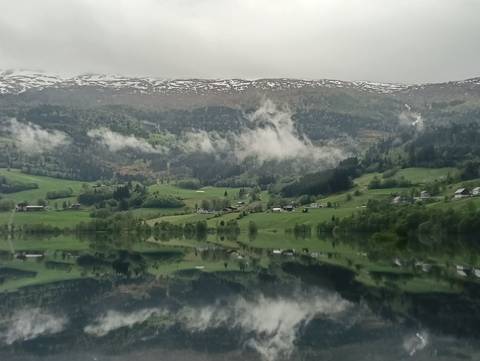 Mirror-calm fjord reflects lush green slopes and low clouds beneath snow-dusted ridge.