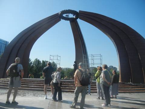 Tour group gathers at flame-shaped war memorial arch under bright blue sky.