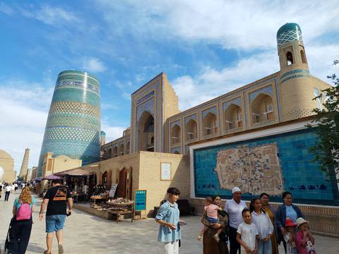 Street scene in an ancient Silk Road city with the turquoise Kalta Minor Minaret and sand-coloured madrasa walls, shoppers and tourists walking under a blue sky.