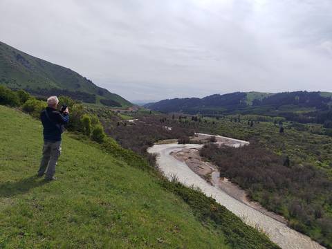 Man taking a photo of a winding river valley surrounded by green hills and distant snow-capped mountains.