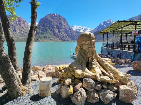 Golden sculpture of a bearded head beside a turquoise lake framed by dramatic snow-tipped mountains.