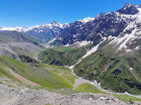 Wide alpine valley with lush green slopes, snow-capped peaks and patches of late-season snow under a clear blue sky.