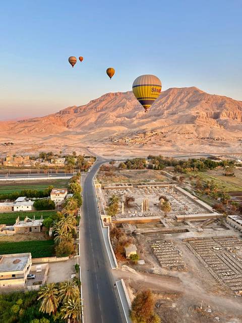 Hot-air balloon floating above desert landscape dotted with ancient ruins and farms in warm sunrise light.