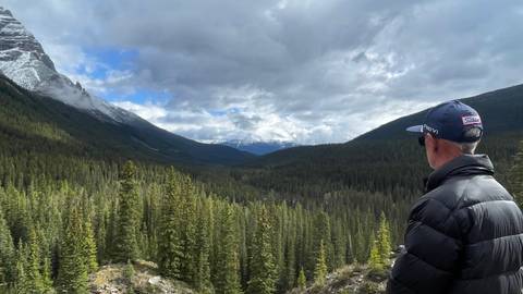 A man in a cap and puffer jacket admires a wide, forest-filled valley framed by snow-dusted peaks beneath a partly cloudy sky.