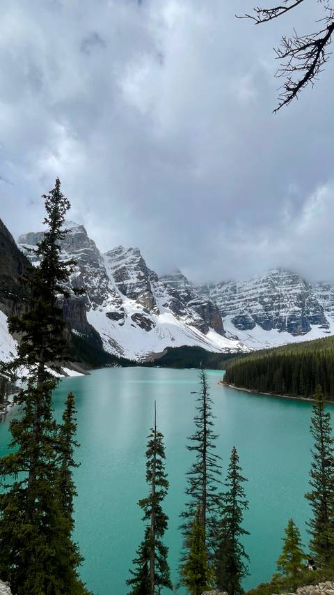 Turquoise glacial lake surrounded by snow-capped jagged peaks and evergreen forests under a dramatic cloud layer.