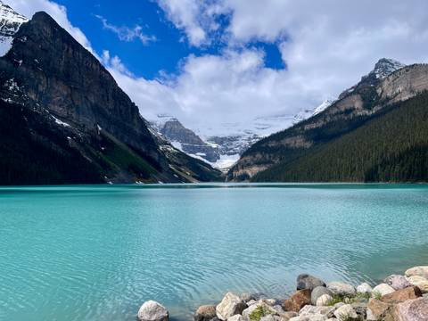 Crystal-blue Lake Louise stretches toward towering cliffs and distant glaciers beneath a patchy blue sky.