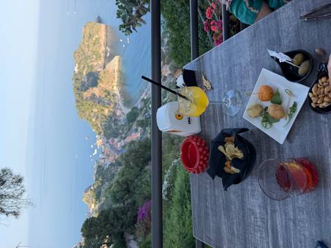 Drinks and Sicilian snacks on a terrace table overlooking a picturesque bay dotted with boats far below.