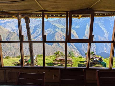 Panoramic mountain scenery framed by a wooden lodge window showing grassy terrace tables high above an Andean valley.