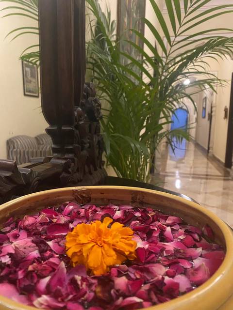 A decorative bowl filled with rose petals sits on a dark table in an elegant hotel hallway lined with green plants and marble flooring.